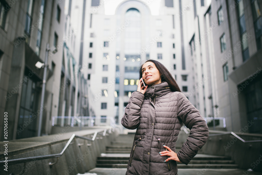 young girl talking on mobile phone in courtyard business center. girl with long dark hair dressed in winter jacket in cold weather speaks on phone on background buildings made of glass and concrete