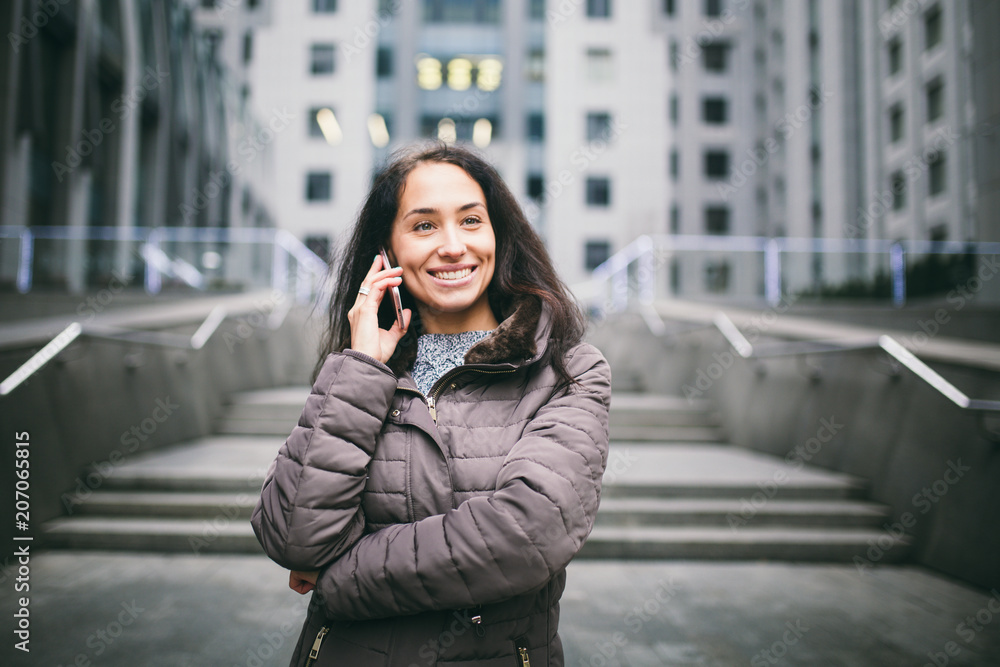 young girl talking on mobile phone in courtyard business center. girl with long dark hair dressed in winter jacket in cold weather speaks on phone on background buildings made of glass and concrete young girl talking on mobile phone in courtyard business center. girl with long dark hair dressed in winter jacket in cold weather speaks on phone on background buildings made of glass and concrete