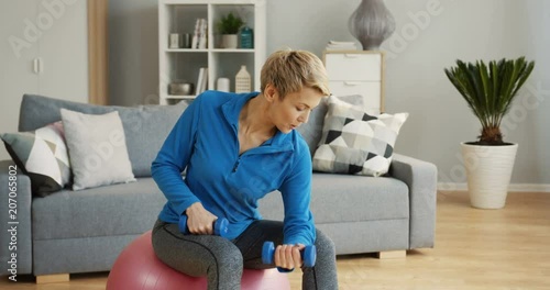 Caucasian middle aged woman in sporty style sitting on the fitness ball and swinging her hands with dumbbells at home. Indoors photo