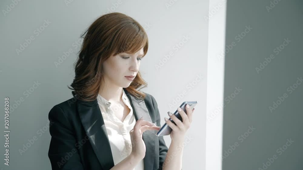 Young attractive business woman is using smartphone standing in bright office, female looking, touching phone screen, chatting during working day indoors. Concept: lifestyle, communication, browsing. Young attractive business woman is using smartphone standing in bright office, female looking, touching phone screen, chatting during working day indoors. Concept: lifestyle, communication, browsing.