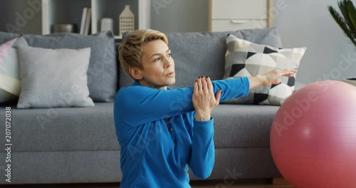 Sporty middle aged woman with short blond hair sitting in the living room and stretching her arms. Indoor photo