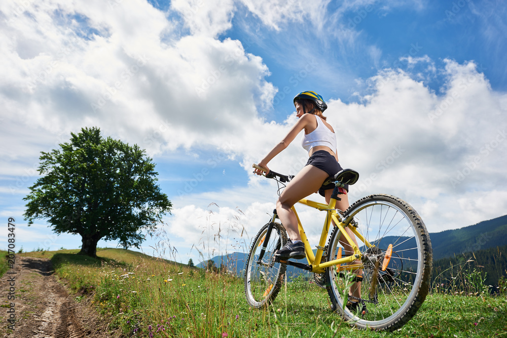 Rear view of sporty female rider cycling on yellow bike on a rural trail in the mountains, wearing helmet, enjoying valley view on summer day. Big tree and cloudy sky on the background