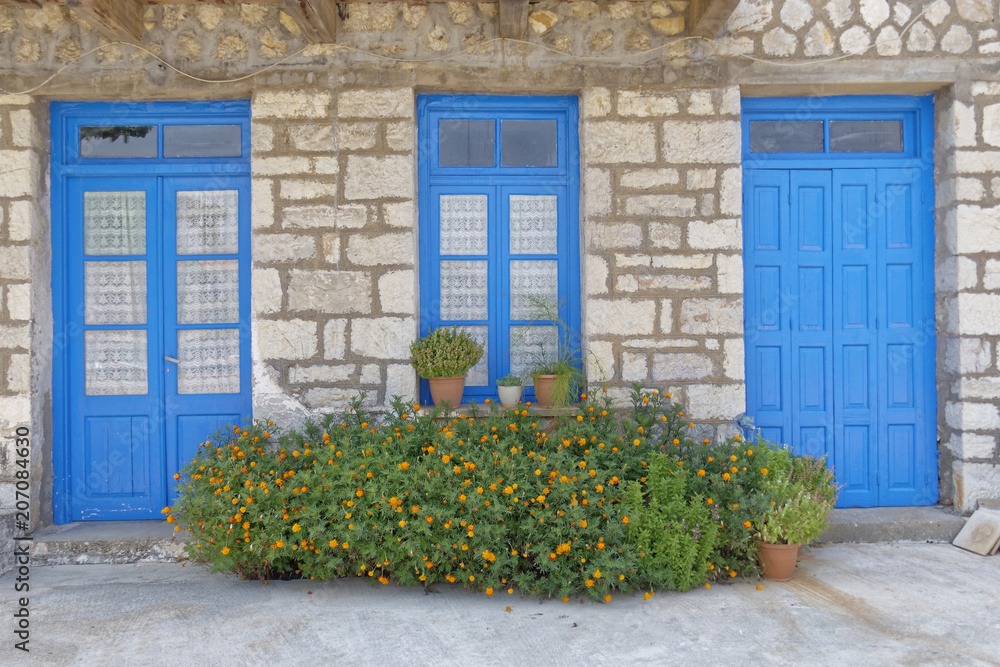 Fototapeta premium Greece, marigold flowers in front of blue doors and window, stone wall house facade