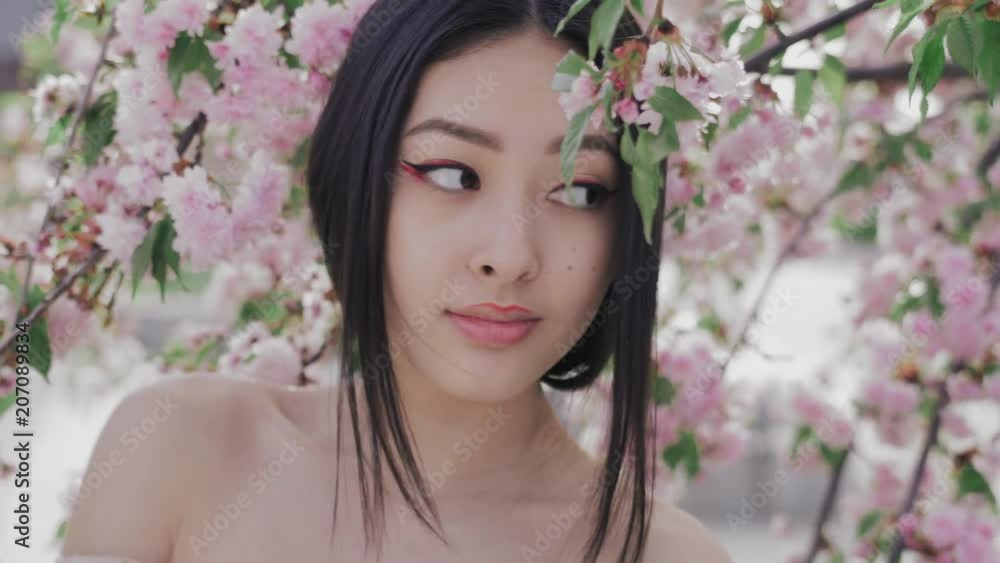 Perfect model with creative vivid makeup and pink lipstick on lips and traditional japanese hairstyle posing outside happy looking at camera. Spring blossom of sakura tree in city park and glamour Perfect model with creative vivid makeup and pink lipstick on lips and traditional japanese hairstyle posing outside happy looking at camera. Spring blossom of sakura tree in city park and glamour