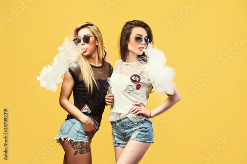 Side view of trendy young female in glasses and shorts standing and smoking on studio background