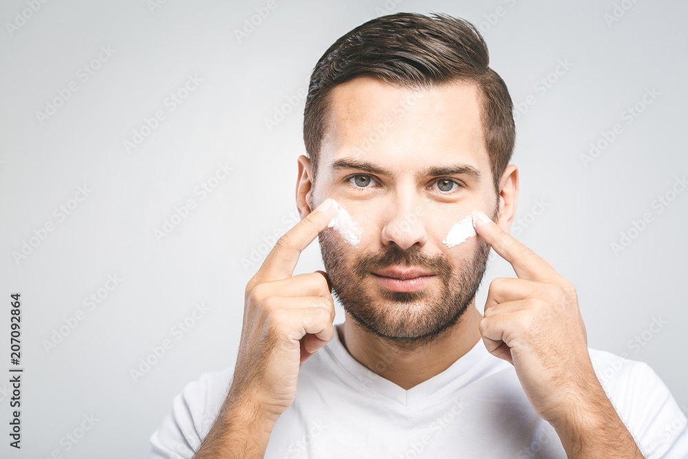 Skin care. Handsome young shirtless man applying cream at his face and looking at himself with smile while standing over gray background and looking at camera. Close-Up. Space for text. Skin care. Handsome young shirtless man applying cream at his face and looking at himself with smile while standing over gray background and looking at camera. Close-Up. Space for text.