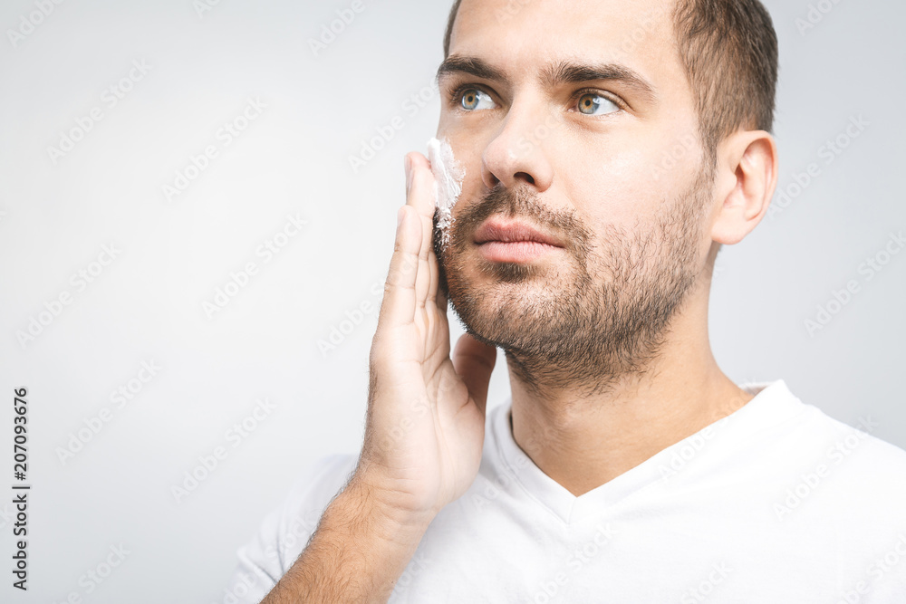 Skin care. Handsome young shirtless man applying cream at his face and looking at himself with smile while standing over gray background. Close-Up. Space for text. Skin care. Handsome young shirtless man applying cream at his face and looking at himself with smile while standing over gray background. Close-Up. Space for text.