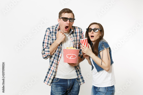 Young shocked couple, woman and man in 3d glasses watching movie film on date, holding bucket of popcorn and plastic cup of soda or cola isolated on white background. Emotions in cinema concept.
