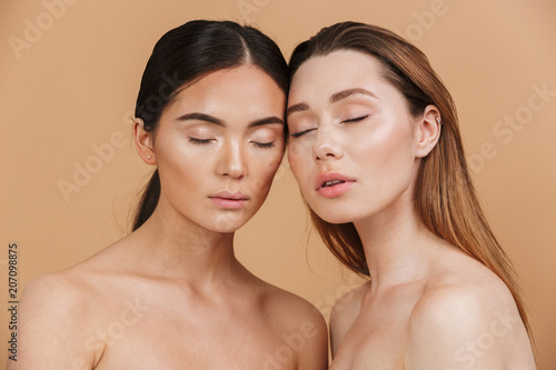 Beauty portrait closeup of two different nation women, asian and caucasian nude girls with closed eyes, posing together at camera isolated over beige background