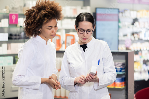 Two experienced female pharmacists wearing lab coats while analyzing together the package of a new pharmaceutical drug in the interior of a modern pharmacy