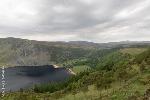 Panoramic view of mountain range, green hills, vivid green color, mountain lake with black water in the foreground, summer, overcast day