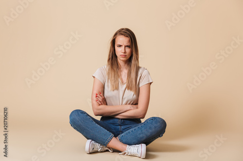 Photo of frowning european woman expressing offense or resentment while sitting on floor with legs and arms crossed, isolated over beige background