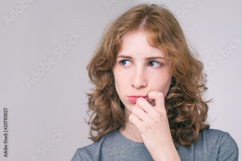Indoor studio portrait of young beautiful redhead European female isolated on white background wearing dress with long curly hair, looking with doubt expression. Toned effect