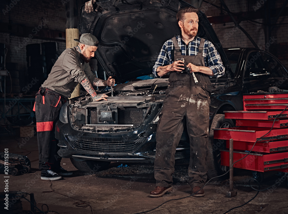 Two bearded auto mechanic in a uniform, repair a broken car in the garage. 