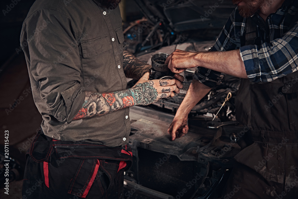 Two mechanics examine a detail of the engine while standing near a broken car in a garage. Two mechanics examine a detail of the engine while standing near a broken car in a garage.
