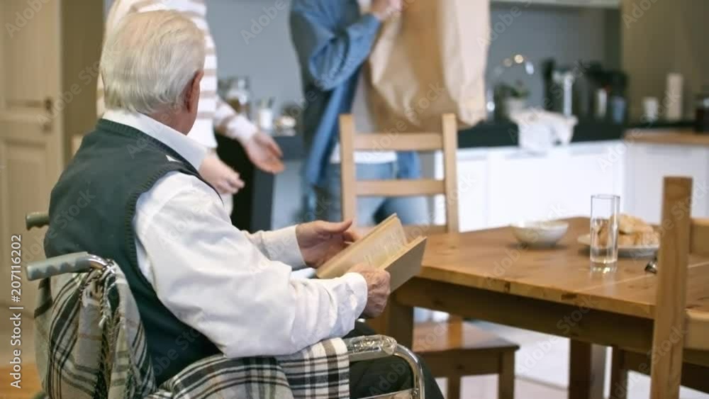 Tilt up of cheerful female caregiver walking into kitchen and chatting with elderly man in wheelchair while male volunteer unpacking groceries Tilt up of cheerful female caregiver walking into kitchen and chatting with elderly man in wheelchair while male volunteer unpacking groceries