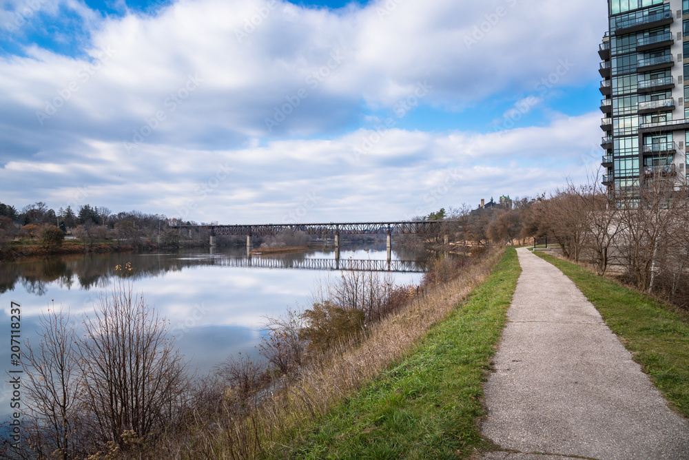 Narrow Path alongside a River and Blue Sky with Clouds on a Late Autumn Day. An Old  Railway Bridge Crossing the River in Visible in Background. Reflection in Water.
