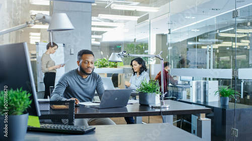 Busy International Office, African-American Man Working at His Desk on a Laptop, in the Background Businesswomen Discuss Relevant Data. Stylish Office with Talented Young People.