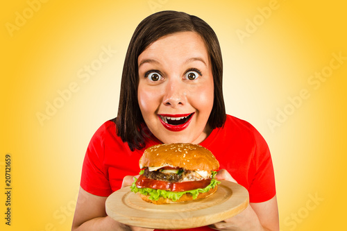 Young woman holds hamburger  smiles and looks in camera. Orange background.