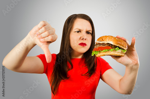 Young unhappy girl holds burger and shows thumb down on gray background.