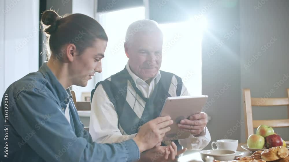 Zoom in shot of happy young man with ponytail showing tablet to grandfather with grey hair and teaching him to use it while having breakfast on sunny morning Zoom in shot of happy young man with ponytail showing tablet to grandfather with grey hair and teaching him to use it while having breakfast on sunny morning