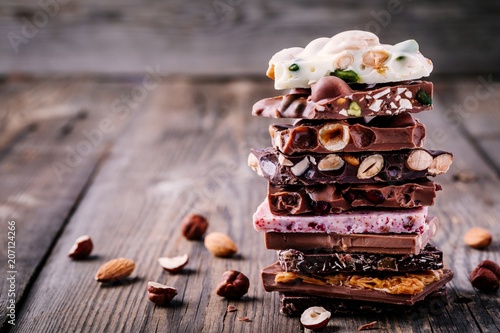 Stack of white, milk and dark chocolate with nuts, caramel and fruits and berries on wooden background.