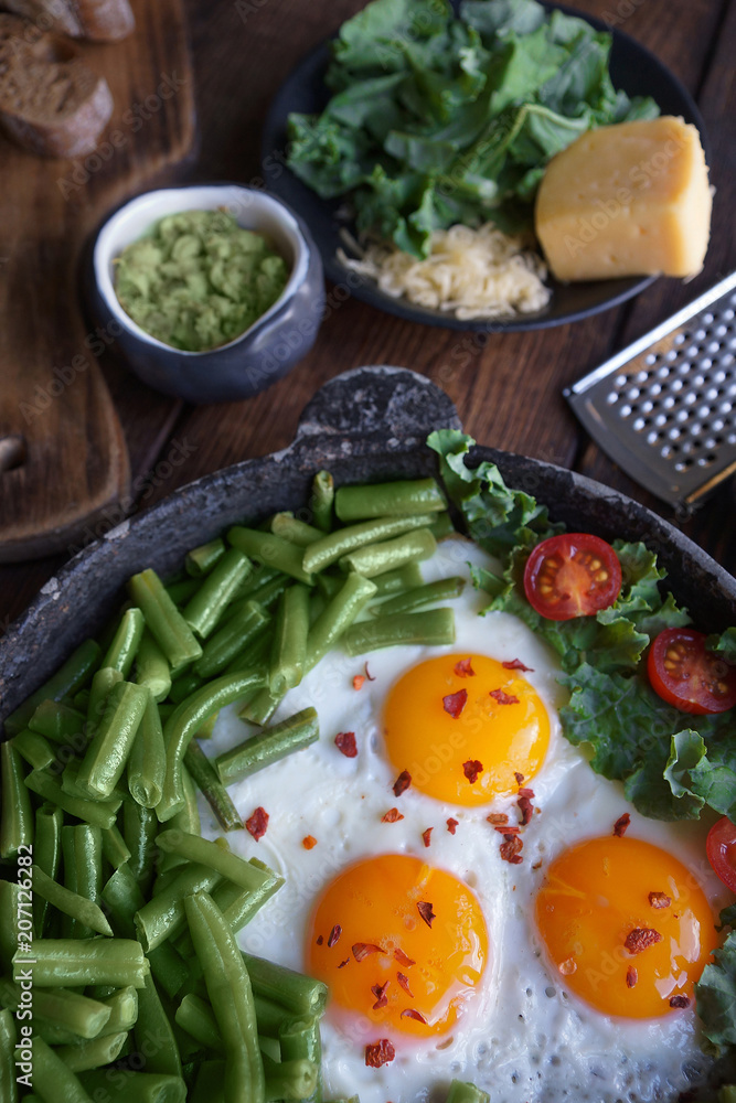 Fried eggs in an old pan with tomatoes, avocado pesto, green beans, pepper and salad on a wooden table, Closeup