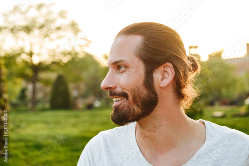 Profile portrait closeup of stylish bearded man 30s with tied hair in white t-shirt smiling, while walking in green park