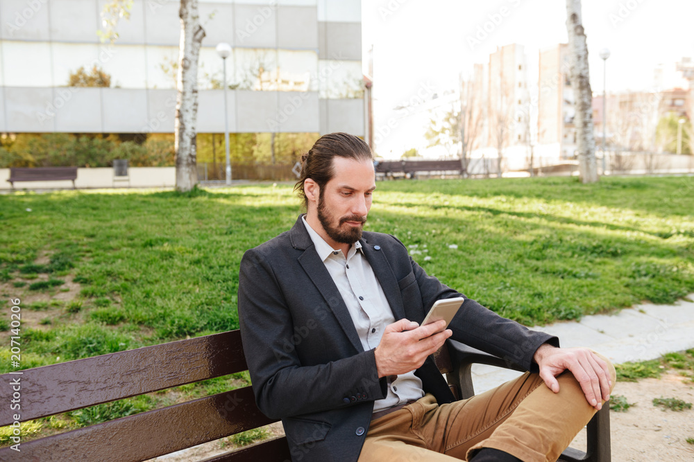 Photo of smart attractive man 30s in businesslike suit holding and using smartphone, while sitting on bench in city park during sunny day Photo of smart attractive man 30s in businesslike suit holding and using smartphone, while sitting on bench in city park during sunny day