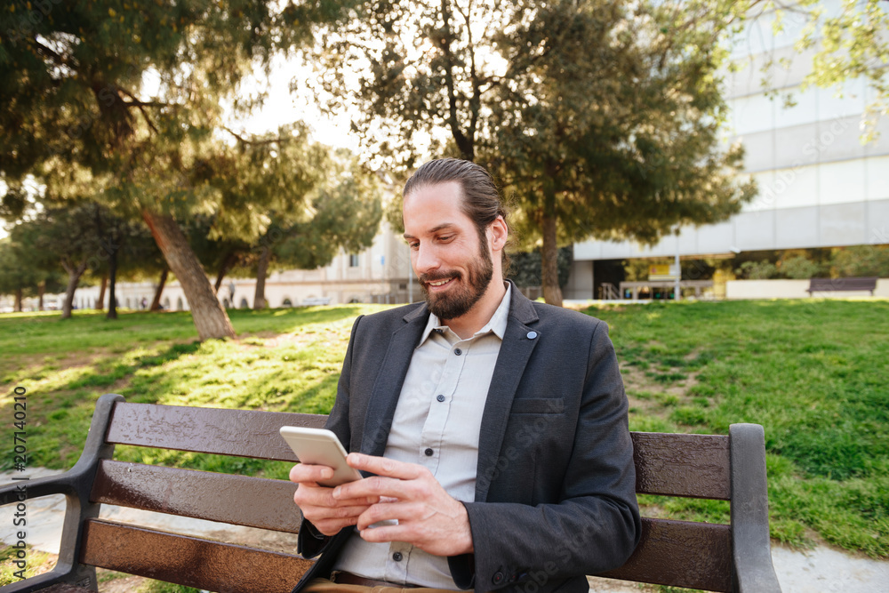 Picture of european bearded man 30s in formal wear holding and typing on cell phone, while resting in city park Picture of european bearded man 30s in formal wear holding and typing on cell phone, while resting in city park