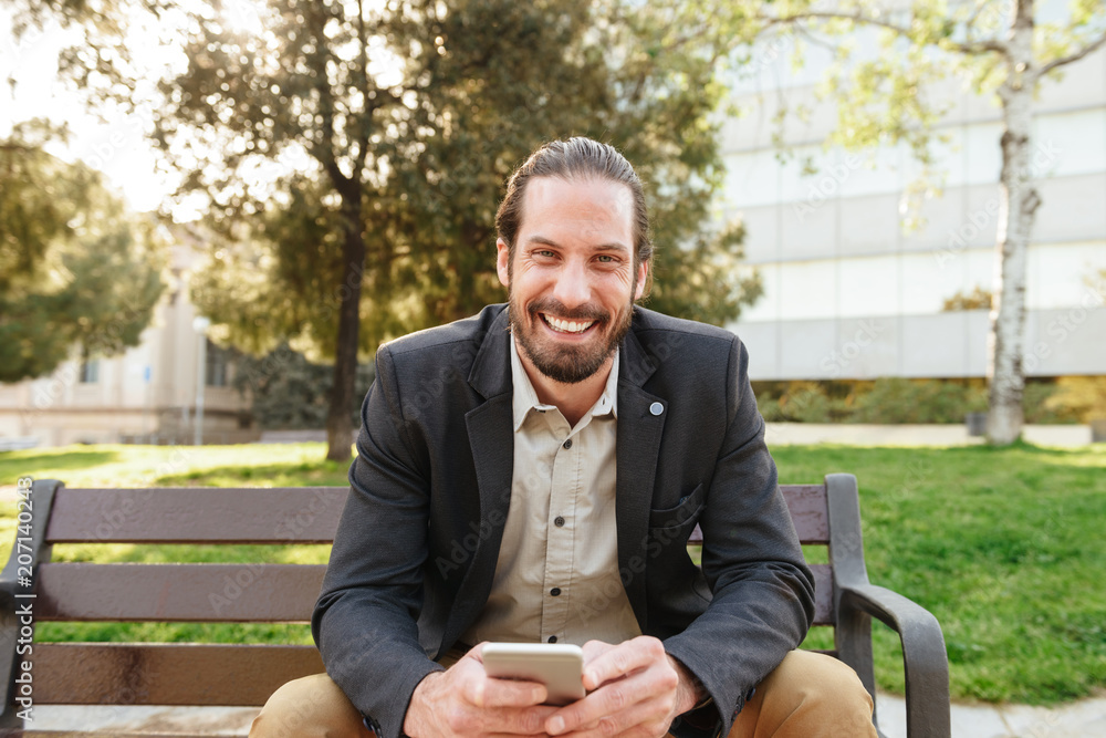 Picture of joyful handsome man with tied hair looking at camera and holding cell phone, while sitting on bench in city park during sunny day Picture of joyful handsome man with tied hair looking at camera and holding cell phone, while sitting on bench in city park during sunny day