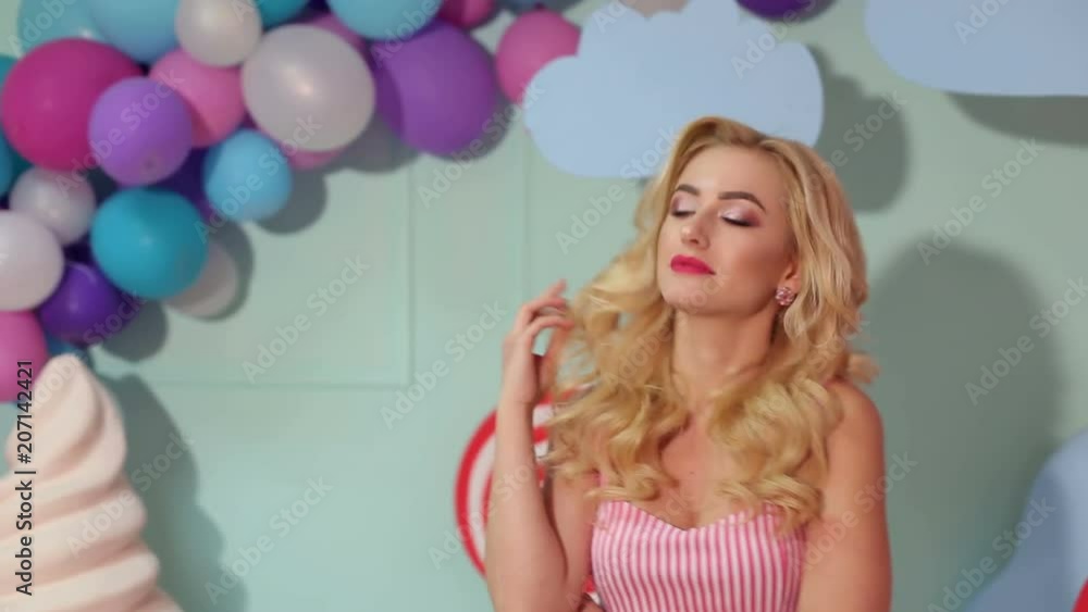 Happy charming girl in a striped pink dress with long blonde hair and bright makeup posing for the camera in the Studio on a colored background. Happy charming girl in a striped pink dress with long blonde hair and bright makeup posing for the camera in the Studio on a colored background.