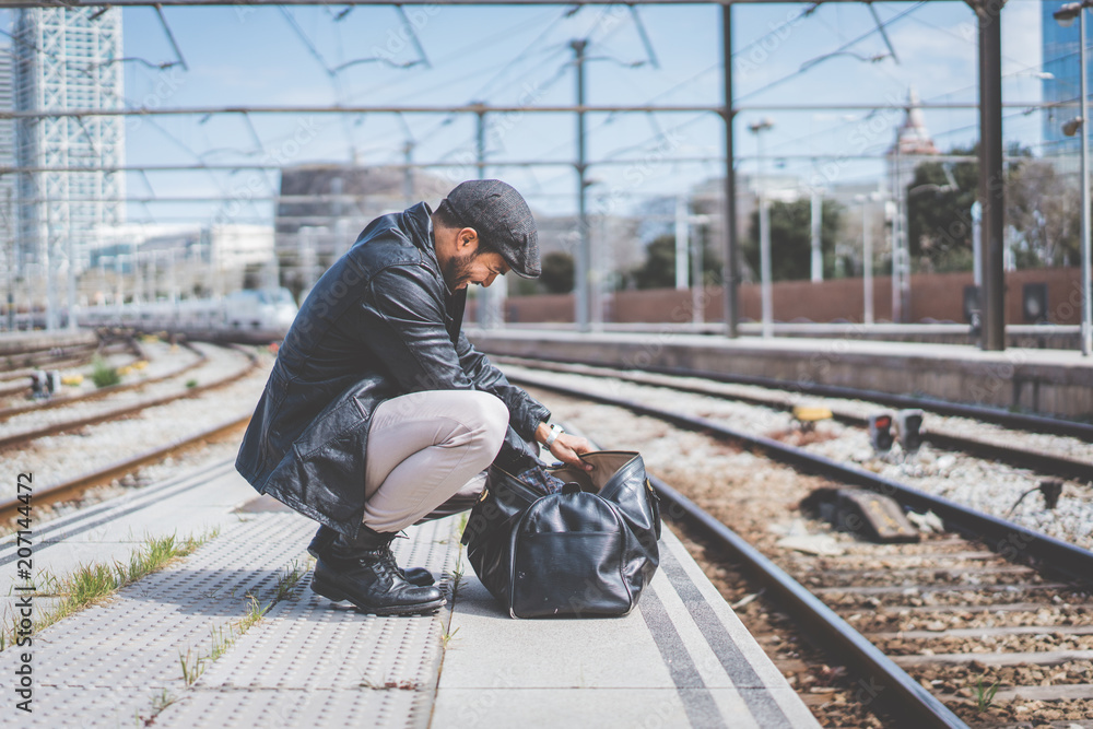 Attractive confident hispanic man having a backpack front of him with a clothes bag, walking beside a platform at train station Attractive confident hispanic man having a backpack front of him with a clothes bag, walking beside a platform at train station