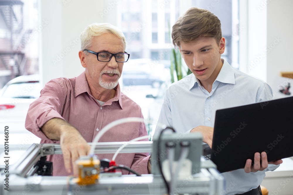 Best teacher. Pleasant senior supervisor teaching his intern about 3D printers while the man holding a laptop