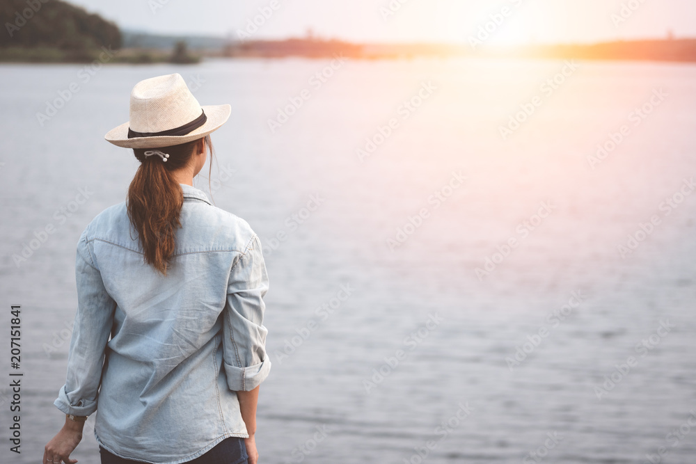Young traveler woman wear denim shirt, jean and hat standing and looking at lake in sunset.