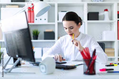 A young girl in the office holds a pen in her mouth and works with a calculator, documents and a computer. photo