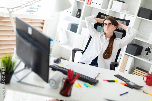 A young girl is sitting at a table in the office, laying her hands behind her head and looking at the monitor.