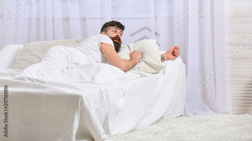Man in shirt laying on bed awake, white curtain on background. Wake up and oversleep concept. Macho with beard and mustache overslept waking up call. Guy on surprised face waking up in morning.