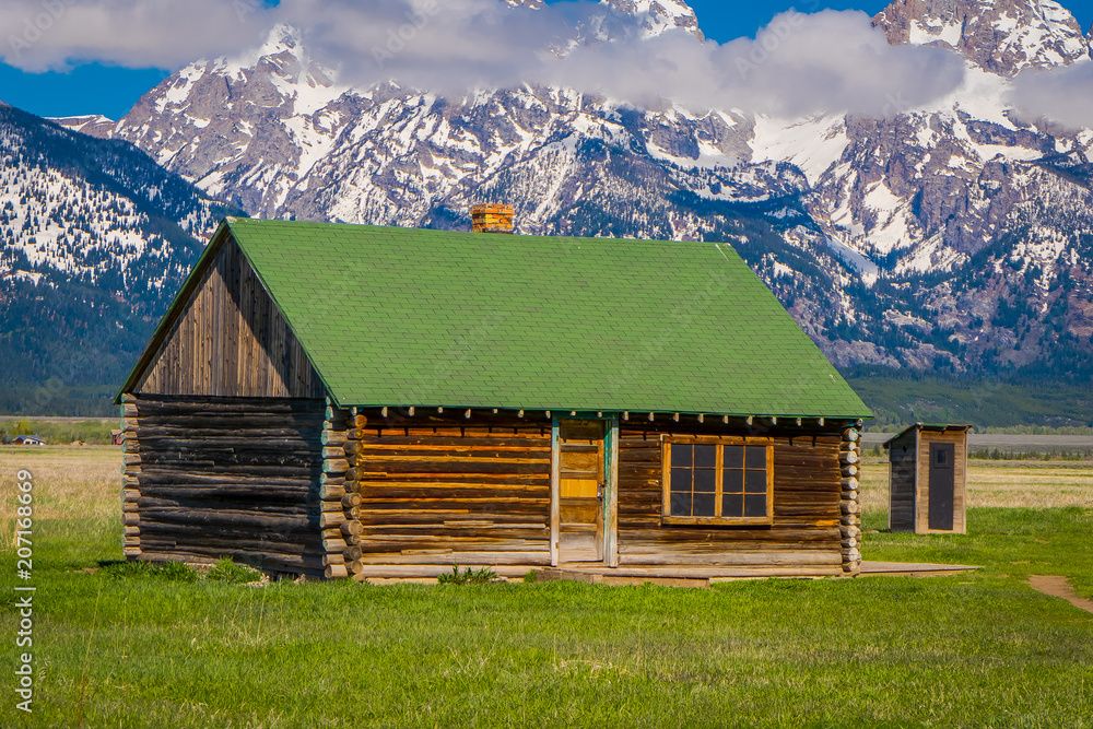 Outdoor view of wooden Cabin with green rooftop on a Golden Grass Prairie against the Grand Teton Mountains, National Park, Wyoming