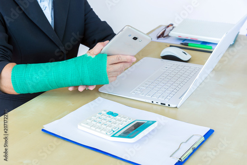 business woman with green cast on arm holding smart phone and working on laptop in office, focus on broken hand