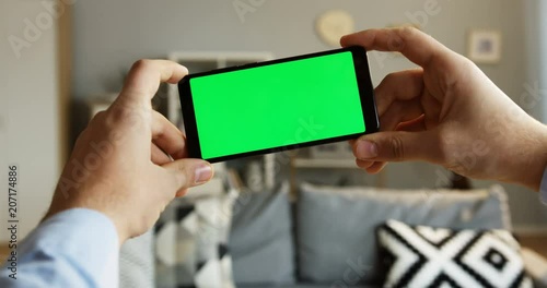 Close up of the male hands holding a black smartphone horizontally with a green screen in the living room. Chroma key. Indoor
