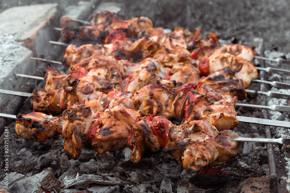 Close up shot of appetizing hot shish kebab with tomatoes on metal skewers prepares on the coals outdoors. Grilling shashlik on barbecue grill.