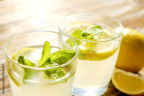 Two glasses full of ice cold refreshing lemonade beverage with mint leaves in sunlight on brown grunged wooden table, slices of ripe organic lemon, whole and halved. Background, copy space, close up.