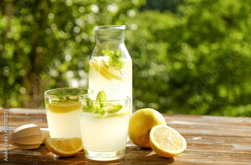 Lemonade pitcher, juicer, two glasses full of ice cold refreshing citrus beverage with lemon slices & mint leaves brown grunged wooden table, country side foliage background. Close up, copy space.
