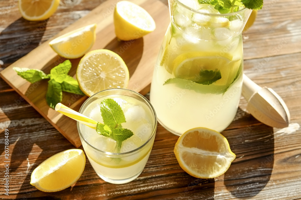 Iced lemonade pitcher, wooden juicer & one glass of cold citrus beverage with lemon slices, mint leaves & yellow straw on brown grunged wood textured table sunny day, background. Close up, copy space. - obrazy, fototapety, plakaty 