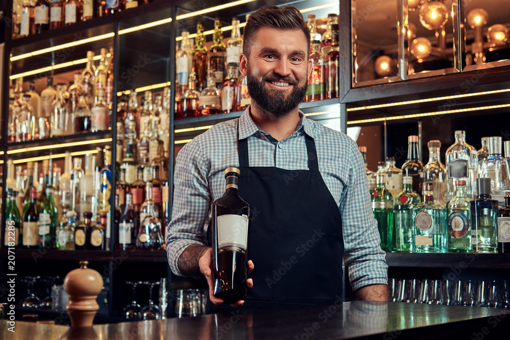 Cheerful stylish brutal bartender in a shirt and apron presents a bottle of exclusive alcohol at bar counter background.