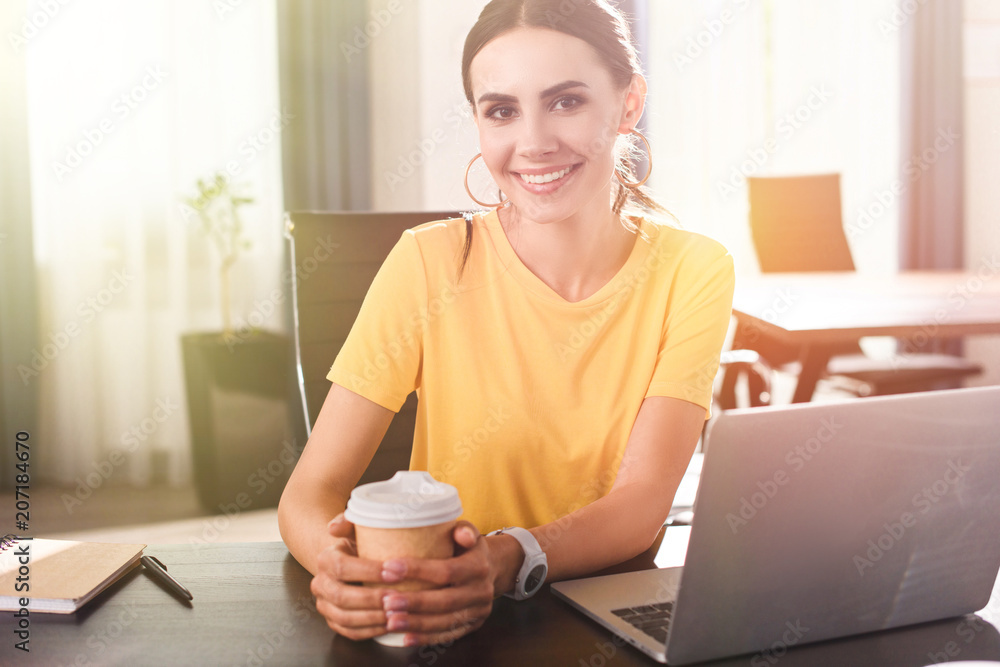 smiling young businesswoman with paper cup of coffee sitting at table with laptop in modern office smiling young businesswoman with paper cup of coffee sitting at table with laptop in modern office