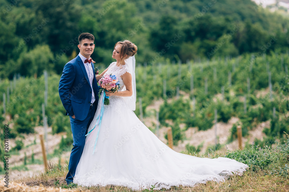 The bride in a blue suit and the bride in a snow-white long dress posing among the greens. The girl is holding a beautiful bouquet, they are happy.