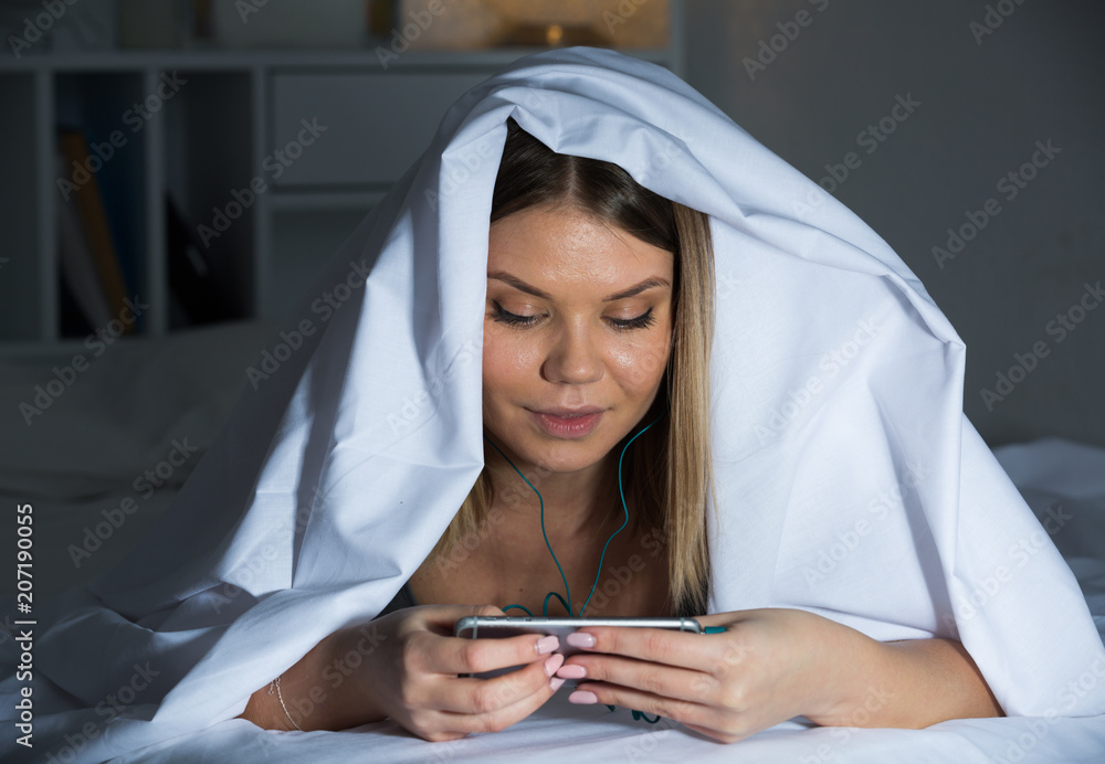 Female under blanket with mobile phone smiling on bed