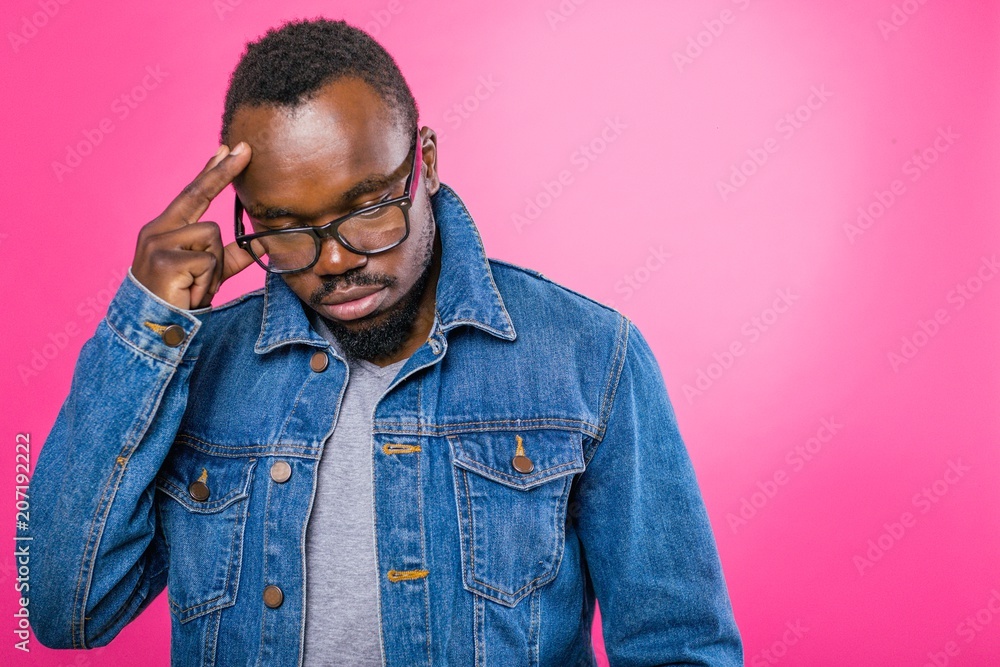 Thoughtful African man in glasses and jeans standing on a pink background thinking raised his hand to his head Thoughtful African man in glasses and jeans standing on a pink background thinking raised his hand to his head